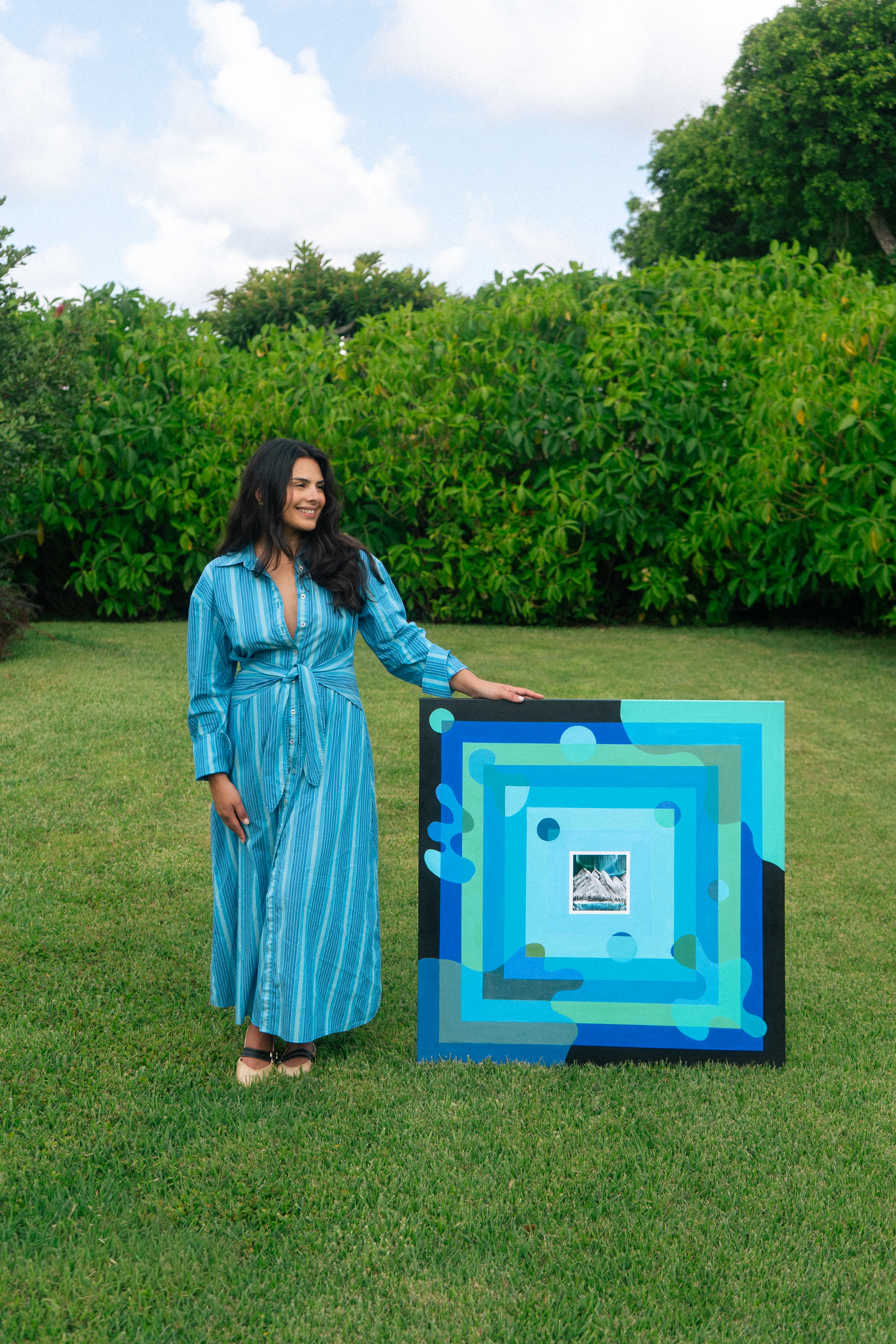 Cristy Bliss in a blue striped dress standing in a lush green garden beside her painting Solo Rider.