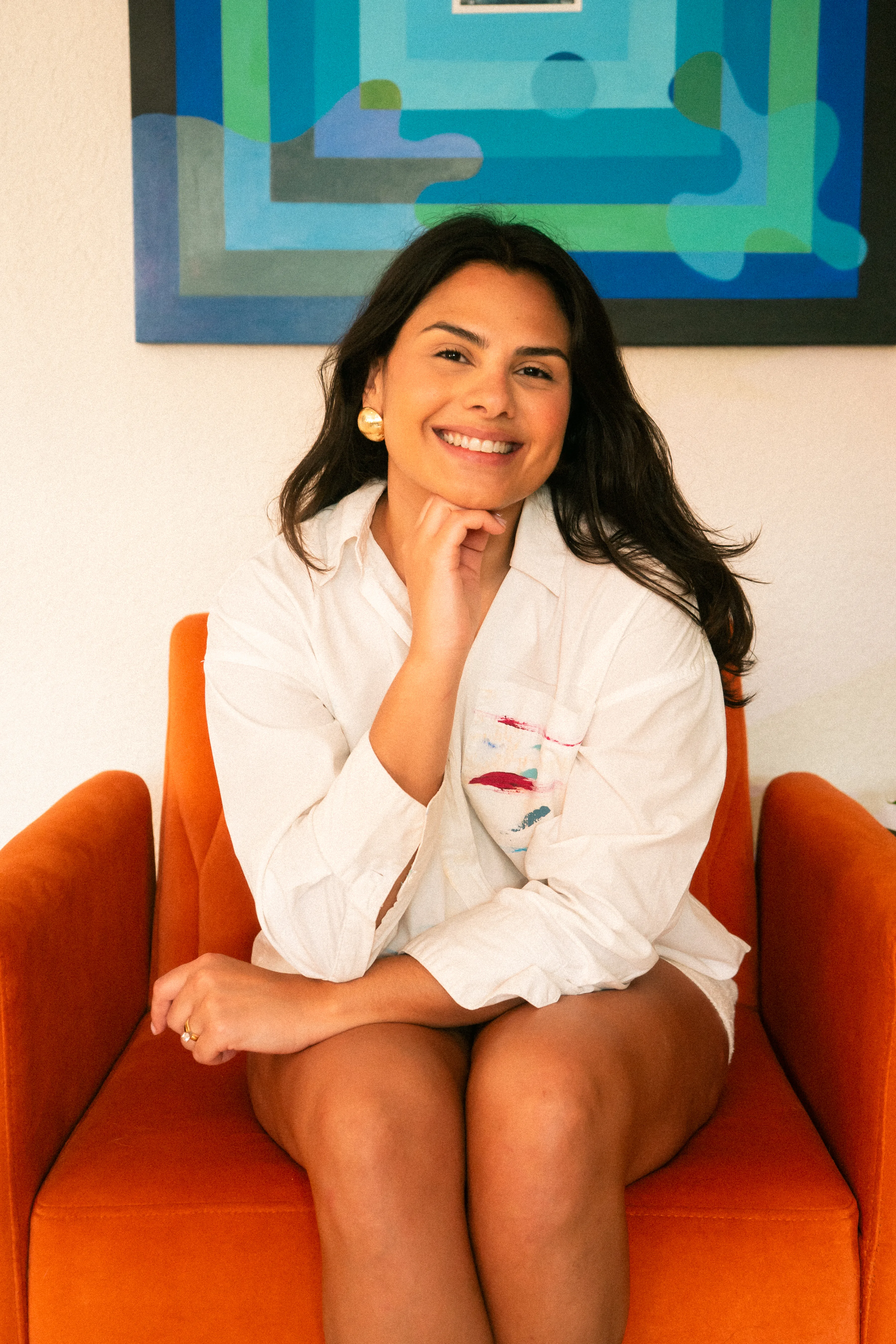 Cristy Bliss seated in an orange chair in a paint-marked white shirt, smiling, one of her blue and green paintings behind her.
