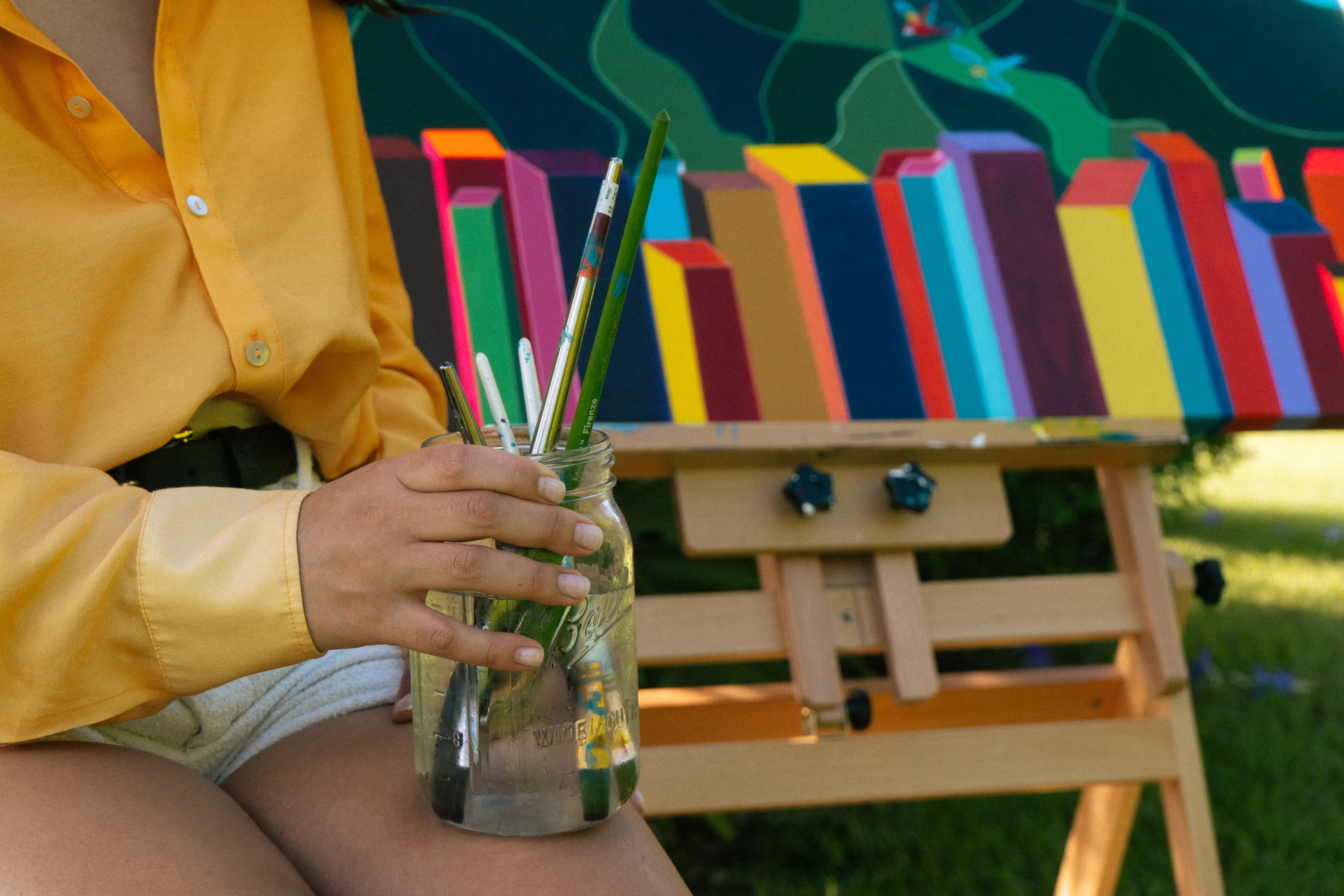 The artist’s hand holding a mason jar of paintbrushes outdoors, her painting Ávila on an easel behind.